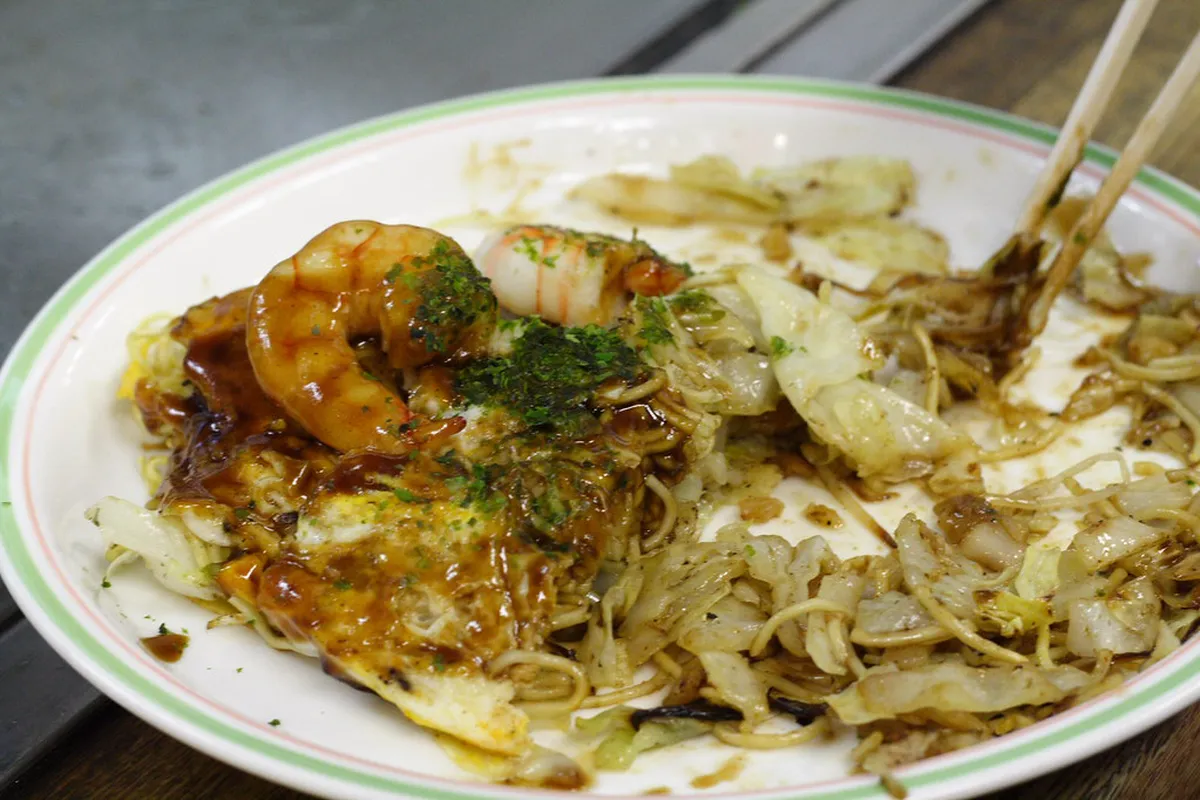A chef cooking a layered Hiroshima-style okonomiyaki on a hot iron griddle with noodles, cabbage, and egg in Hiroshima, Japan
